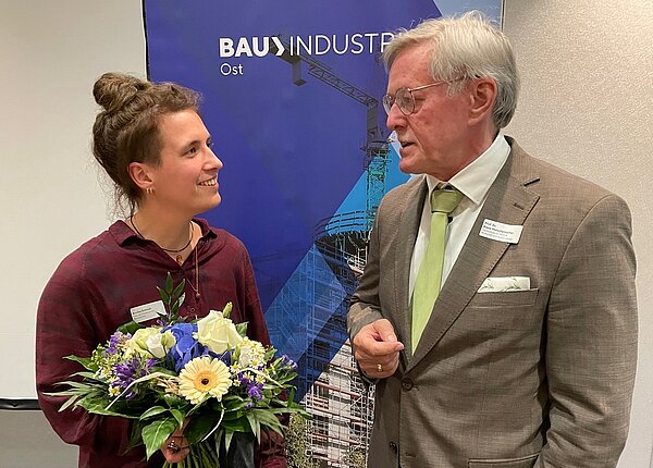 Baupreis 2024 links steht eine Frau und rechts ein Mann, sie schauen sich an. Die Frau hält einen Blumenstrauß.
