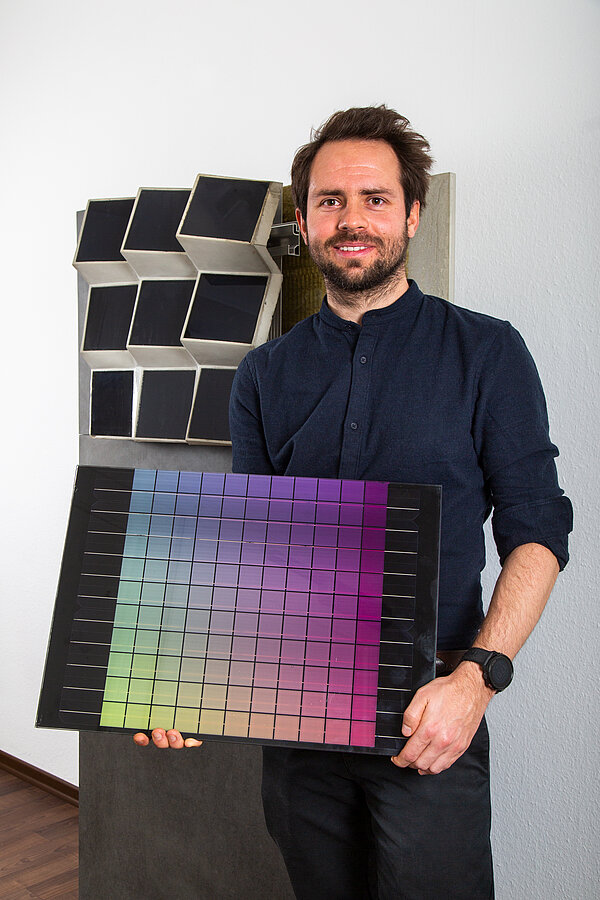 farbige Photovoltaik Adrian Heller hält ein Solarmodul in der Hand, bei dem die Farben von gelb bis rosa verlaufen. Er steht vor einem Modell der Solarcon